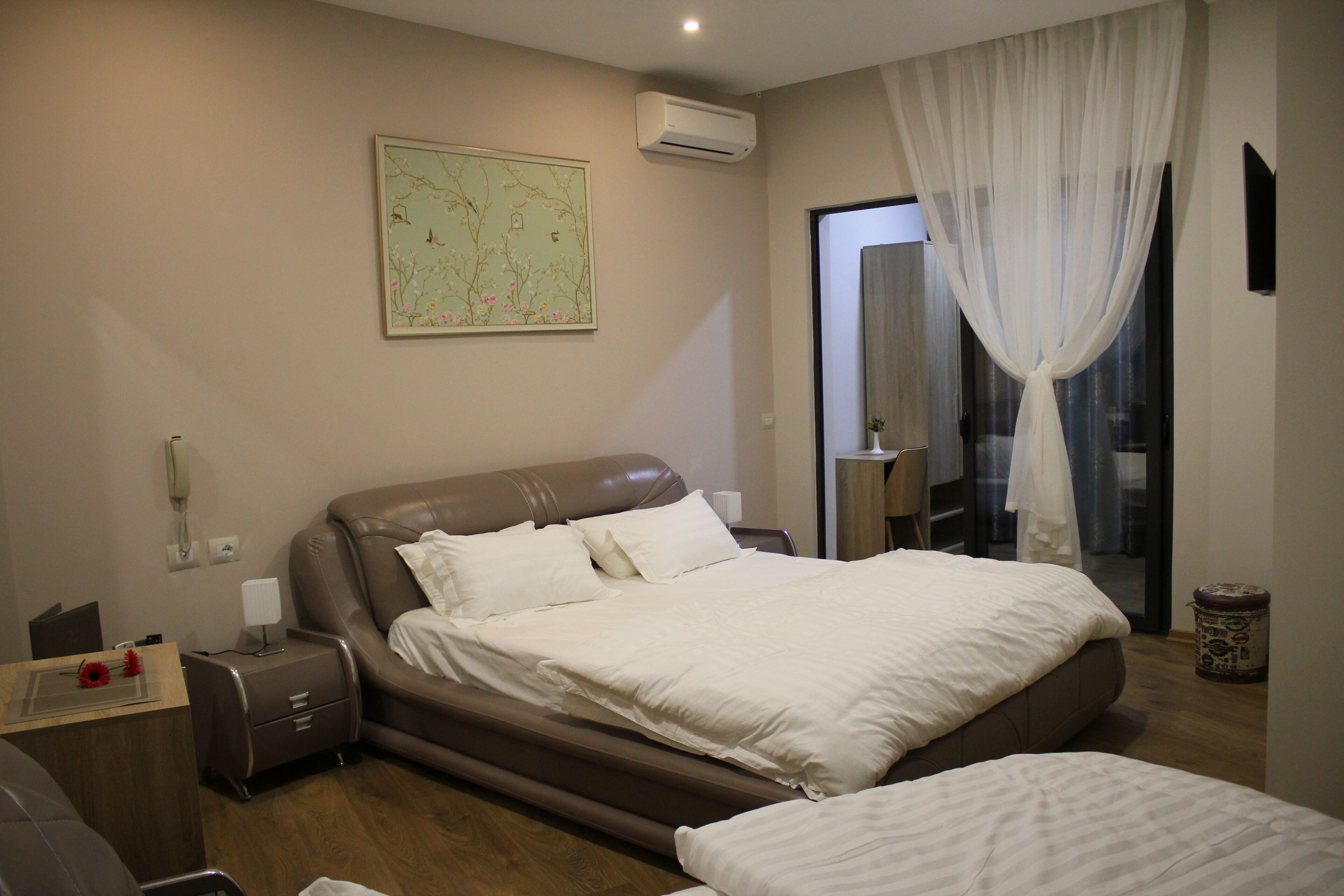 Un dormitorio con una cama grande, una mesita de noche, una lámpara y un mapa en la pared, además de una ventana con cortinas traslúcidas.