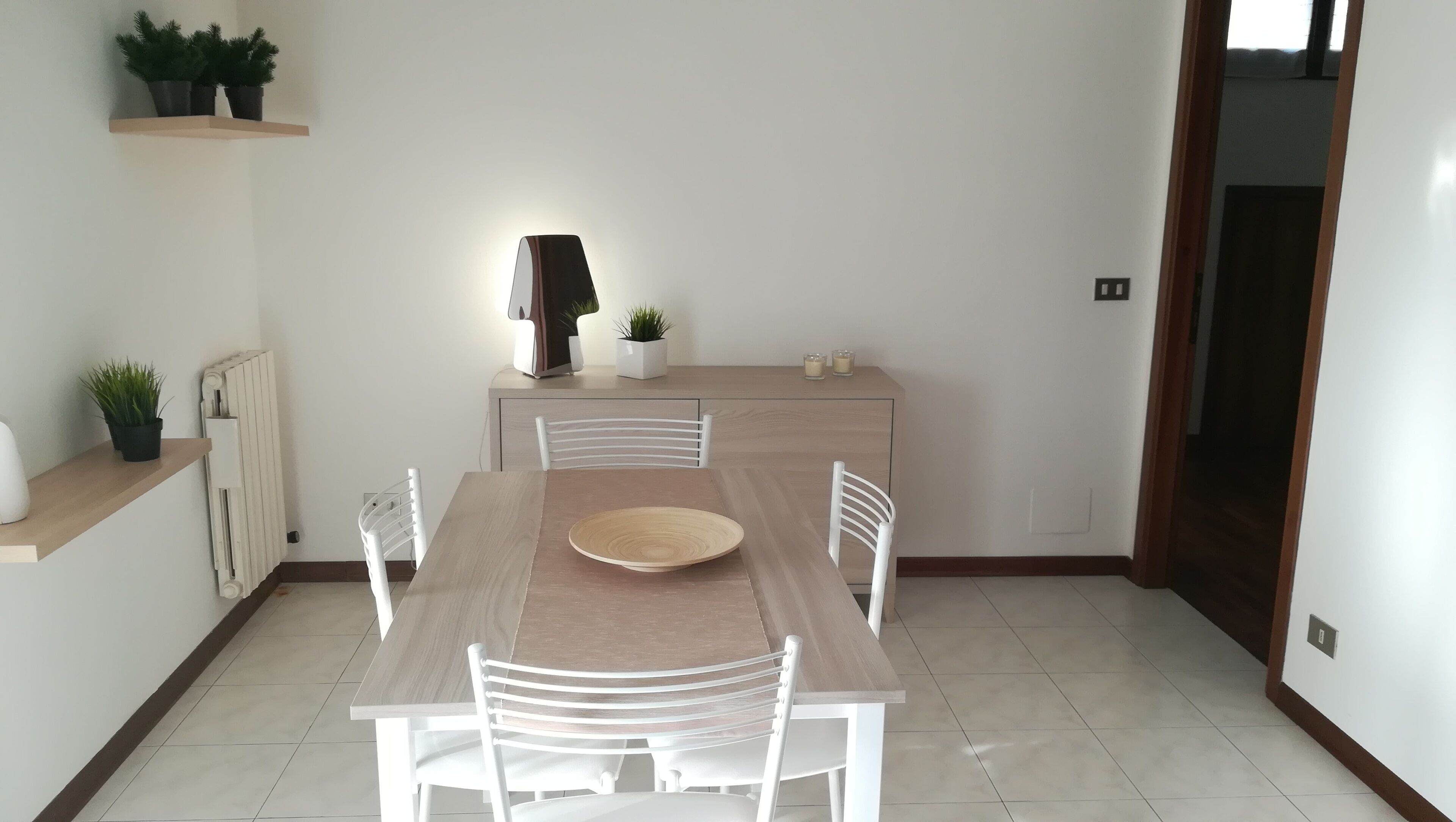A dining area with a wooden table and white chairs, a radiator, and potted plants on a shelf.