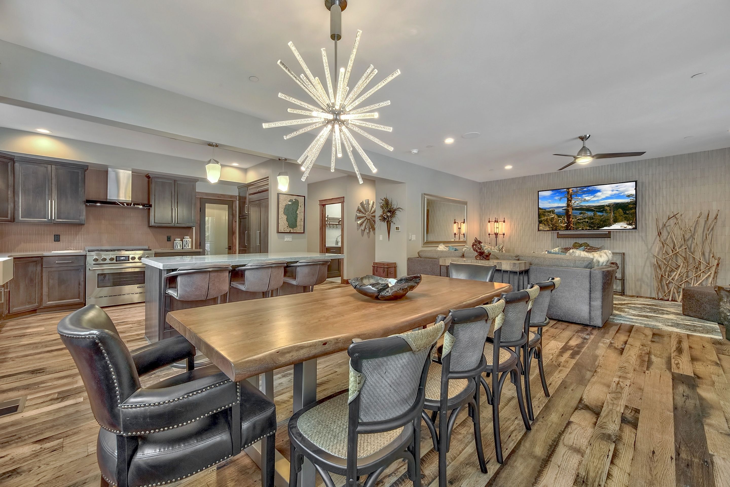 A modern kitchen with stainless steel appliances, a dining area with a wooden table and leather chairs, and a large TV mounted on the wall.