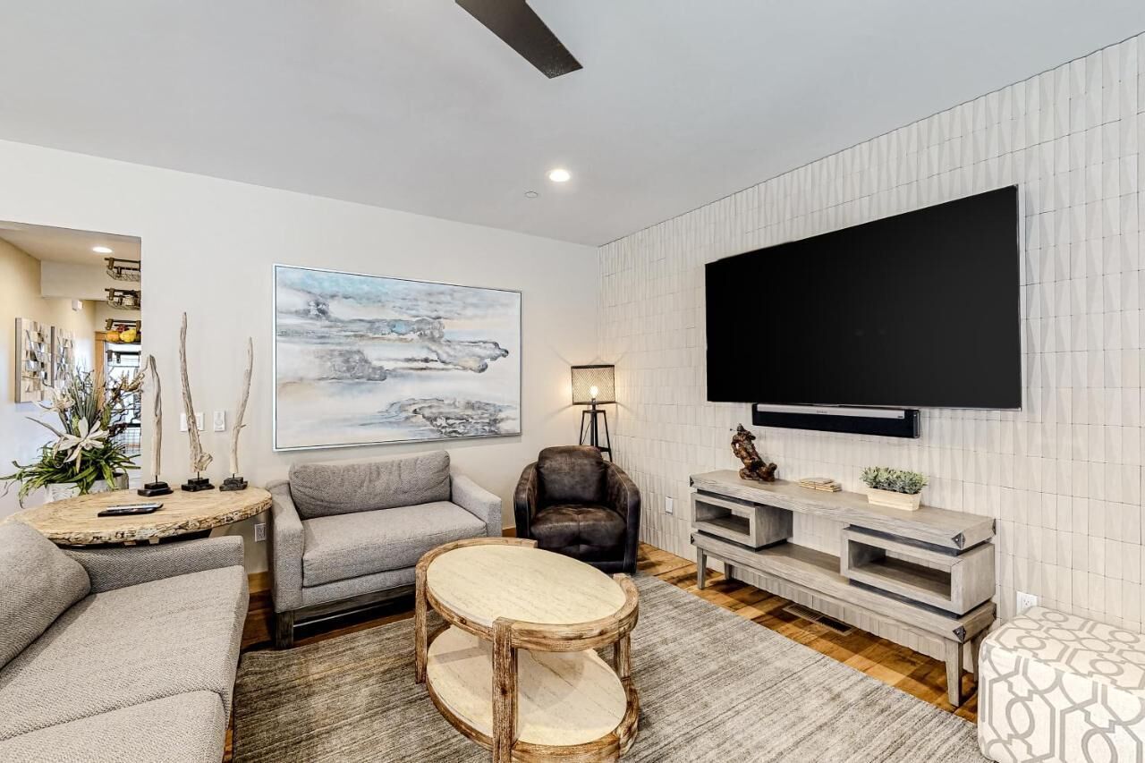 Una sala moderna con un sofá gris, una mesa de centro redonda, un sillón de cuero negro, un televisor de pantalla plana montado en la pared y un mueble bajo de madera con una planta.