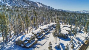 Un complejo turístico nevado con varias cabañas dispuestas en fila, rodeado de un bosque denso y montañas.