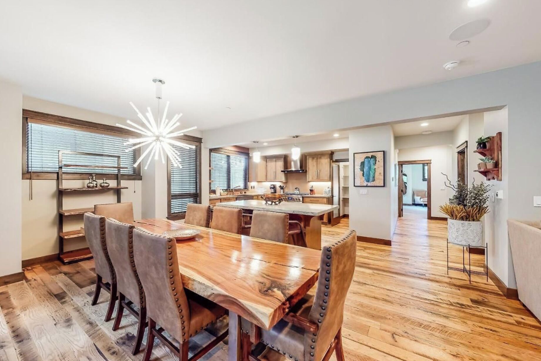 A spacious dining area with a wooden table and chairs, a modern kitchen in the background, and a large window with blinds.