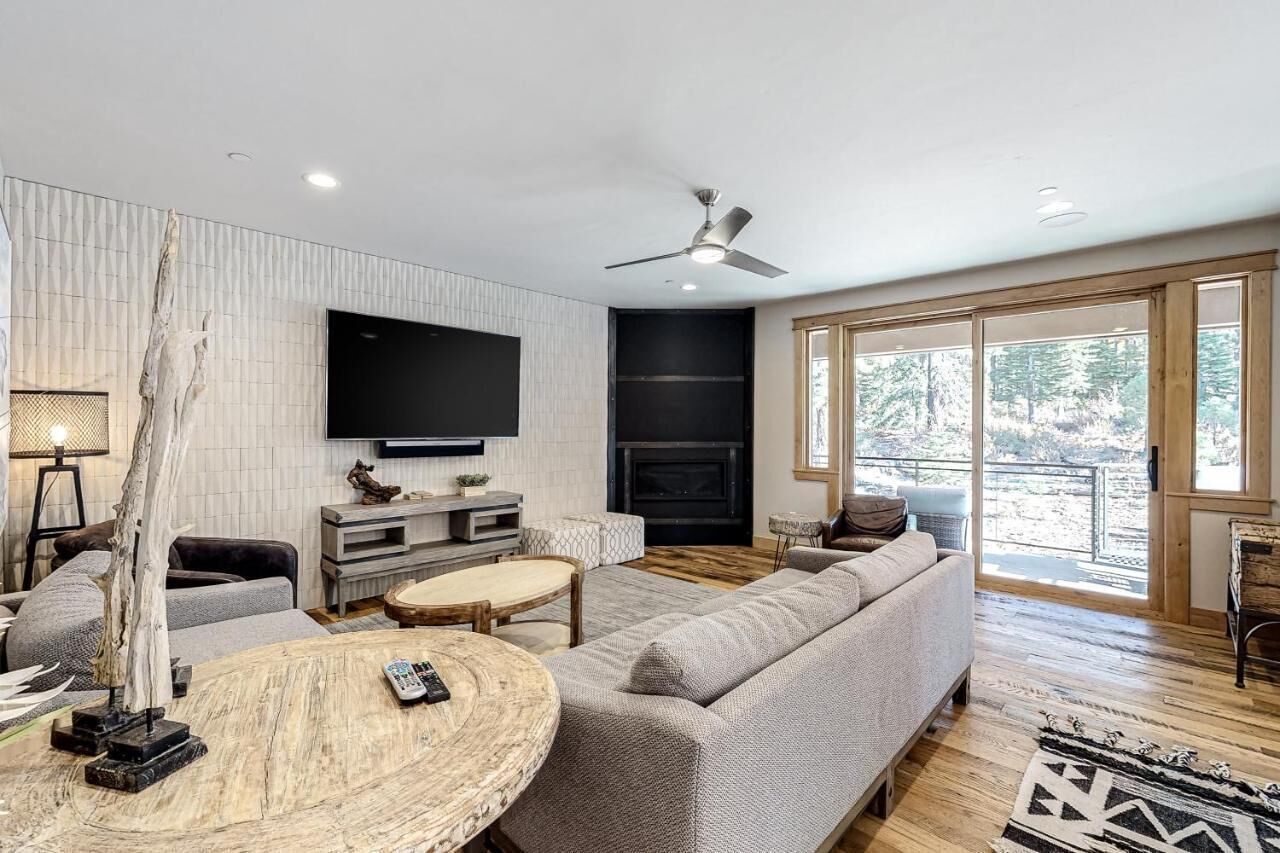 Een moderne woonkamer met een grote flatscreen televisie, een open haard, een plafondventilator en een groot raam met uitzicht op bomen.