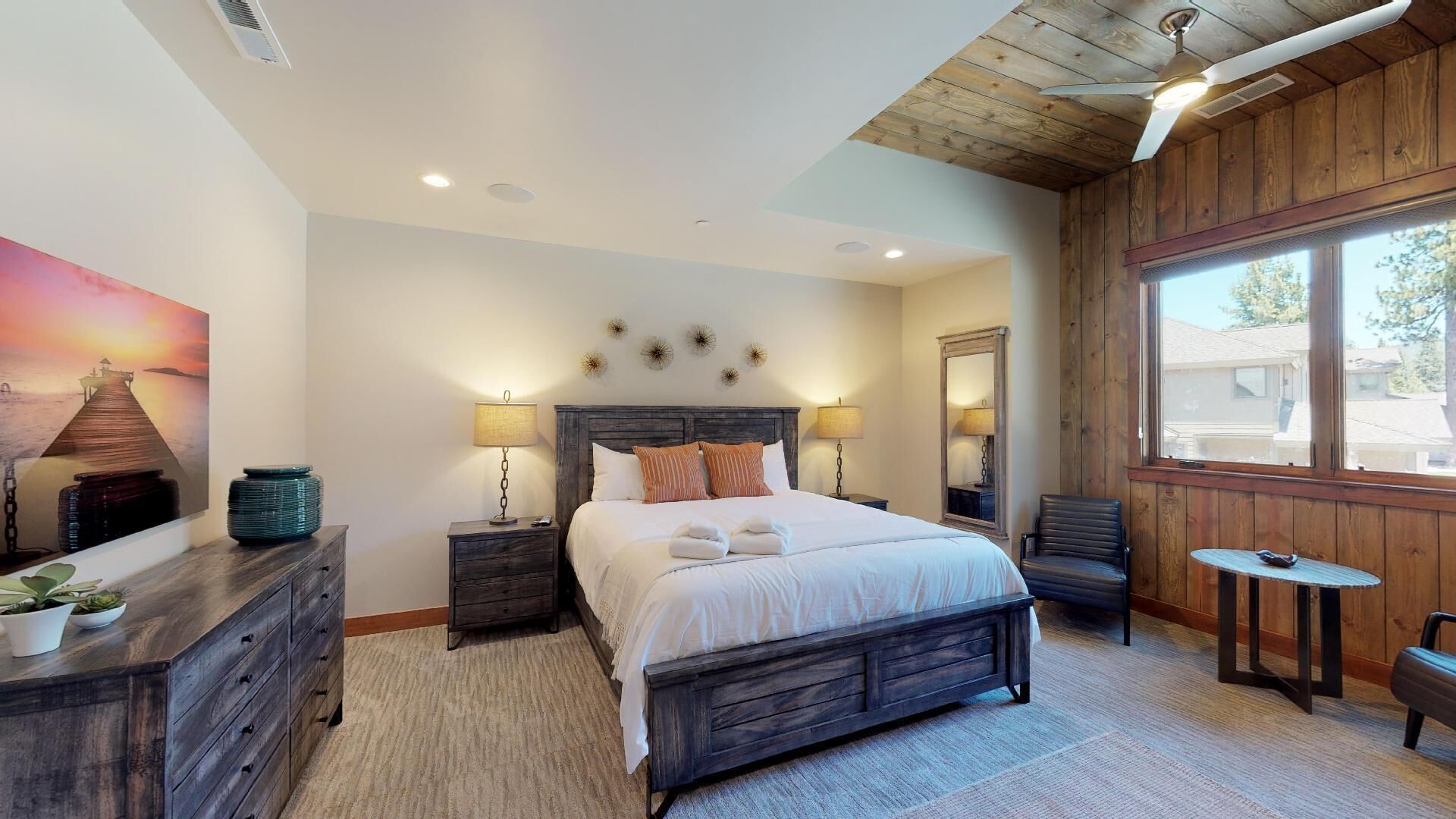 A bedroom with a wooden bed, bedside tables with lamps, a dresser, a chair, a small table, and a large window.