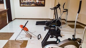 Een kamer met fitnessapparatuur, waaronder een hometrainer, elliptische trainer en een stoel. Op de muur hangt een schilderij van een stadsgezicht.