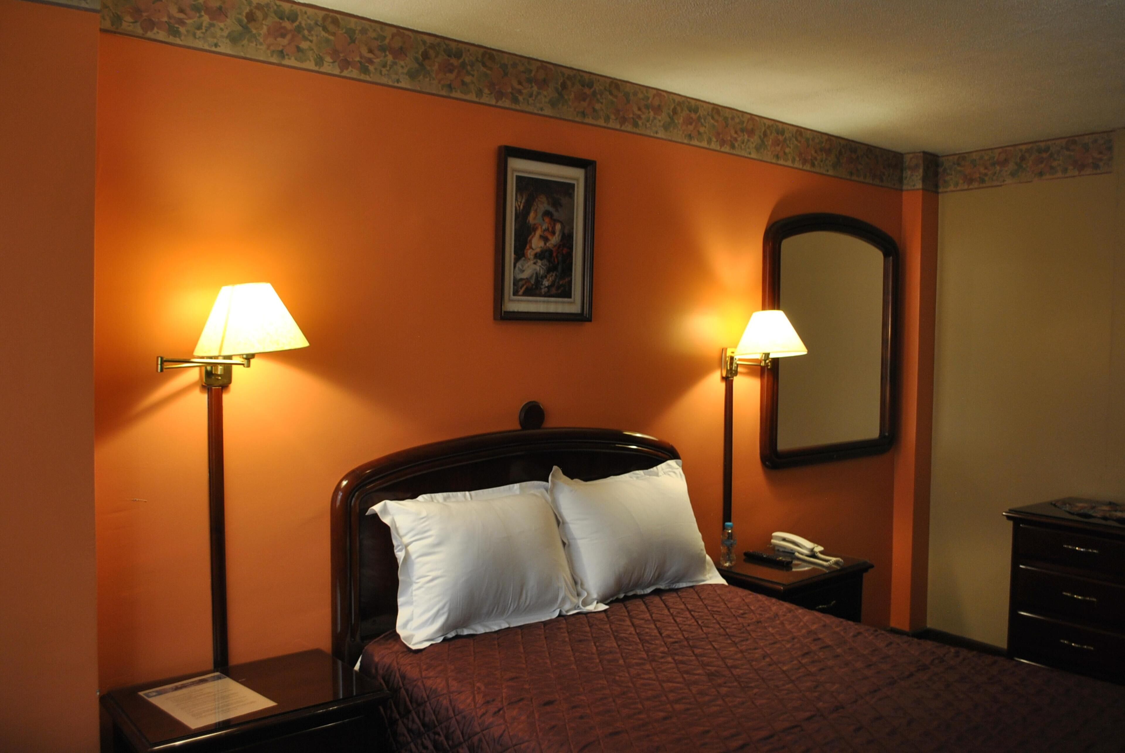 Una habitación de hotel con cama, dos lámparas de noche, una mesita de noche, un espejo y un aparador.