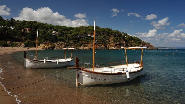 Dos barcos amarrados en una playa con aguas transparentes y un paisaje montañoso al fondo.