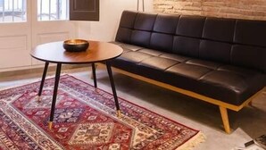 Una sala de estar acogedora con un sofá de cuero oscuro, una mesa de centro redonda de madera, un tapete estampado, un espejo grande y una puerta que da a un balcón.