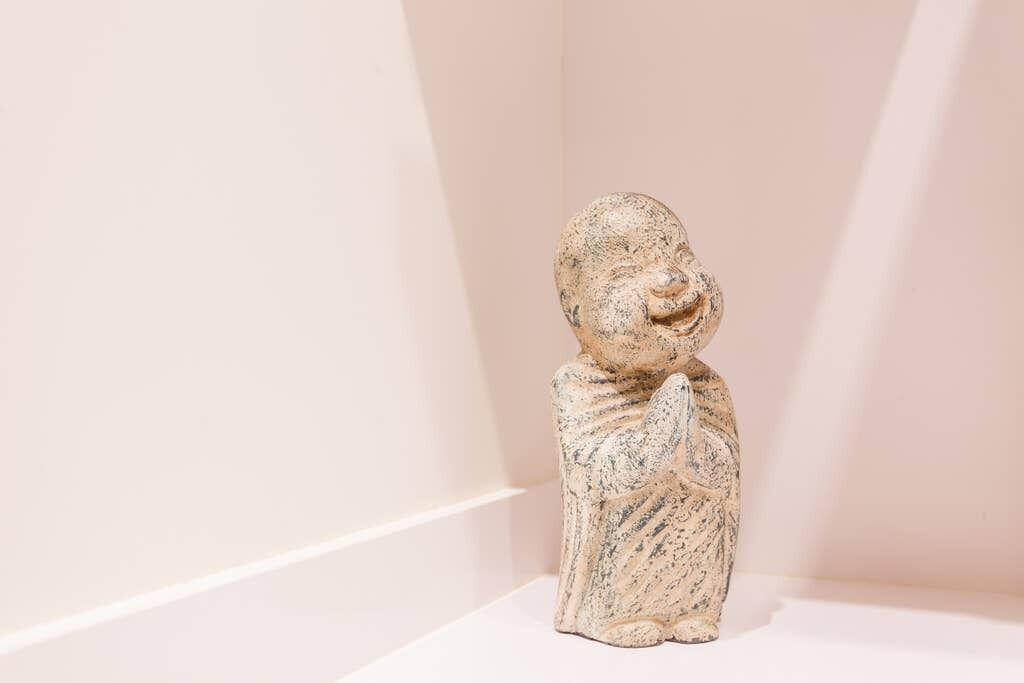 A statue of a seated figure with a rounded head and a small nose, positioned against a plain wall.