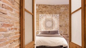 Un estrecho pasillo que conduce a un dormitorio con pared de piedra, cama con ropa de cama blanca y marrón, y un separador decorativo de metal.