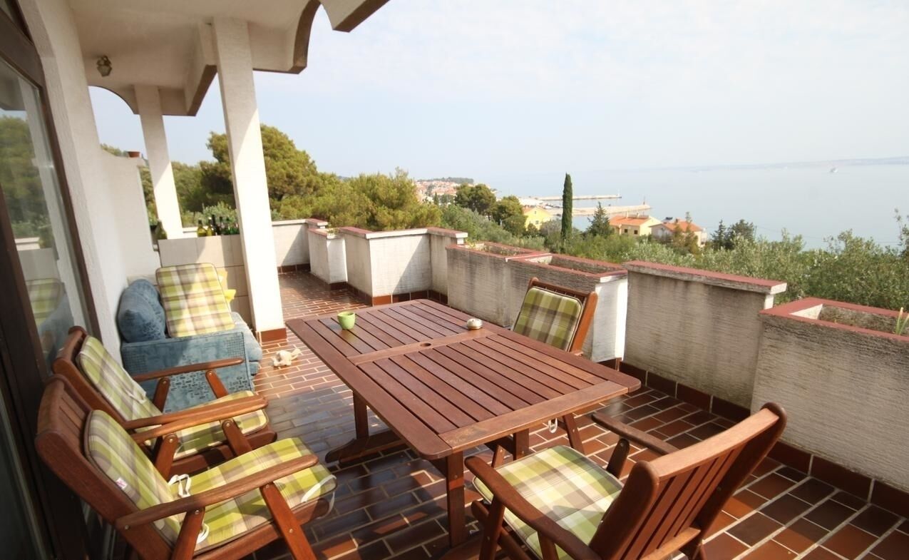 An outdoor patio with a wooden table and chairs, a sofa, and a view of the sea and hills.