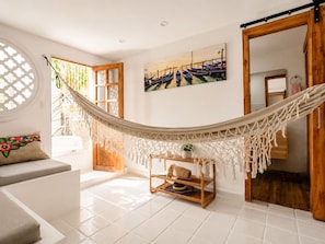 Una habitación con una hamaca, un estante de madera con sombreros, un cuadro y una ventana que da a vista de barcos.