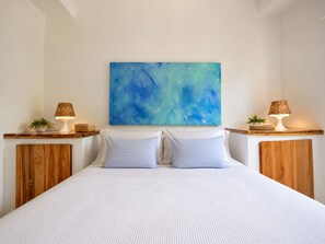 Een slaapkamer met een groot bed, twee nachtkastjes met lampen, een blauw abstract schilderij aan de muur en een houten hoofdbord.