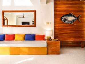 Un dormitorio con cama, almohadas coloridas, cabecera de madera, espejo montado en la pared, ventilador de techo y decoración con peces.