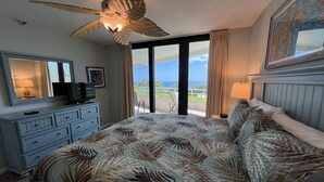 A hotel room with a bed, a ceiling fan, a TV, a dresser, and a large window with a view of the ocean.