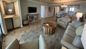 A spacious living room with a large sofa, a coffee table, and a ceiling fan. There is a kitchen area with a dining table and chairs.