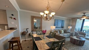 A dining area with a wooden table and chairs, a chandelier, and a view of the living room with a sofa and a large window.