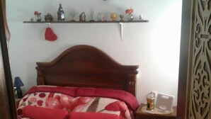 Une chambre à coucher avec une tête de lit en bois, une couvre-lit à motifs floraux, une table de chevet avec une lampe et une étagère ornée de divers objets décoratifs.