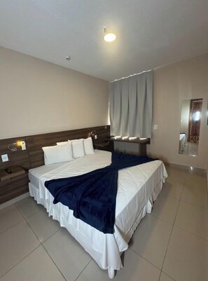 A hotel room with a large bed, a bedside table, a nightstand, a wall-mounted lamp, and a window with curtains. - Hotel Santos Dumont Aeroporto SLZ (São Luís)