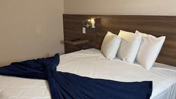 Habitación de hotel con cama, almohadas, manta, mesita de noche y lámpara de pared.