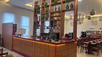 A hotel lobby with a reception desk, wooden shelving with decorative items, and a dining area with tables and chairs.