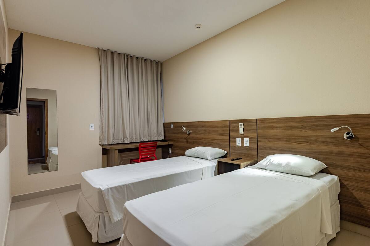 Une chambre d’hôtel avec deux lits simples, une tête de lit en bois, un petit bureau, une chaise rouge et un miroir.