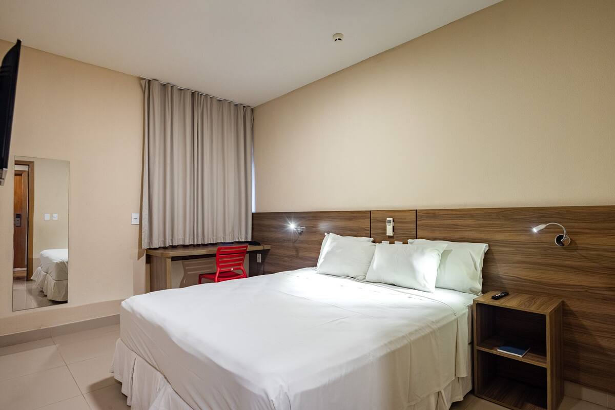 Une chambre d’hôtel avec un grand lit, un bureau avec une chaise, un miroir et une table de chevet avec une lampe.