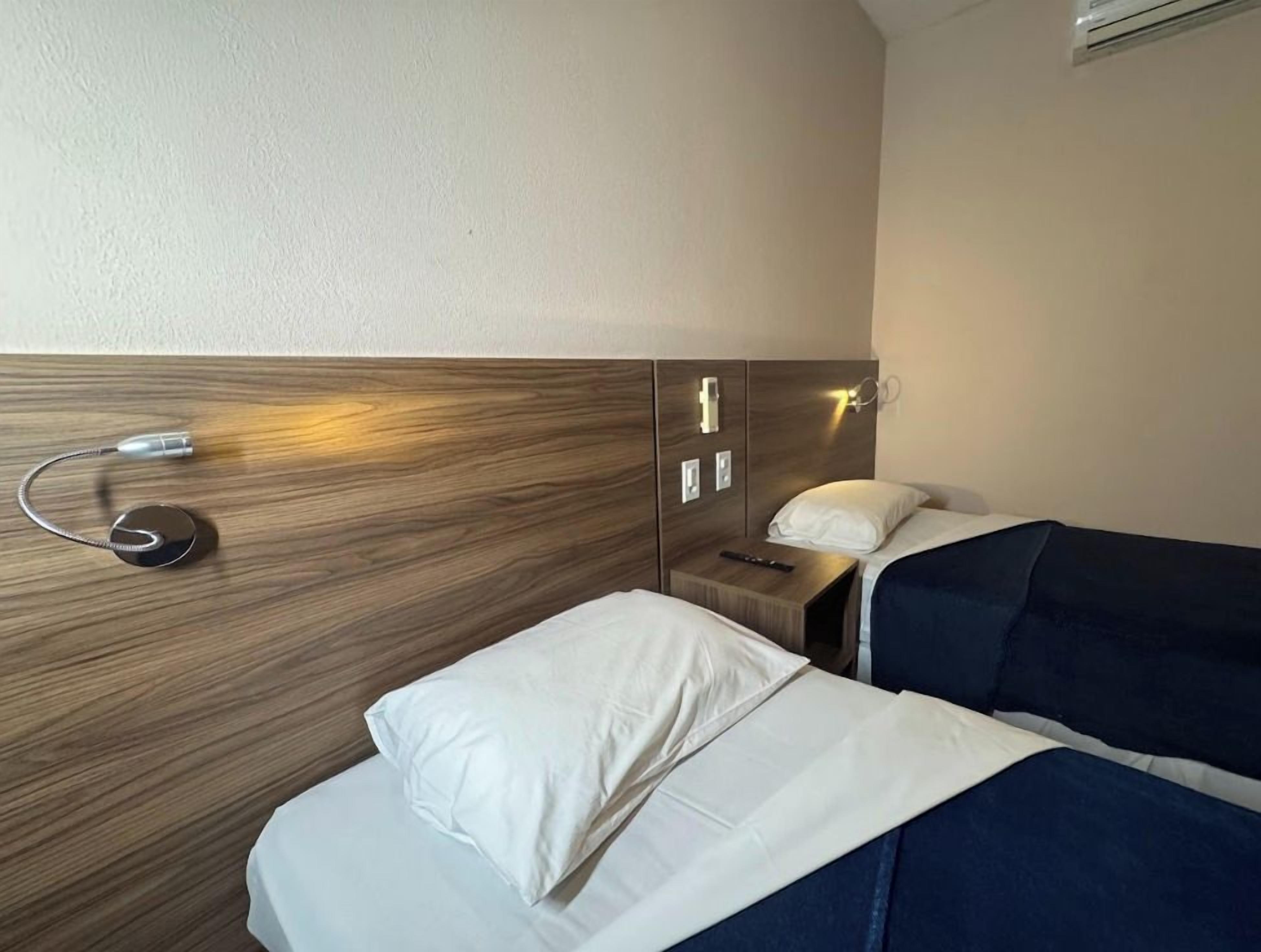 Quarto de hotel com duas camas, uma mesa de cabeceira, abajur e ar condicionado fixado na parede.