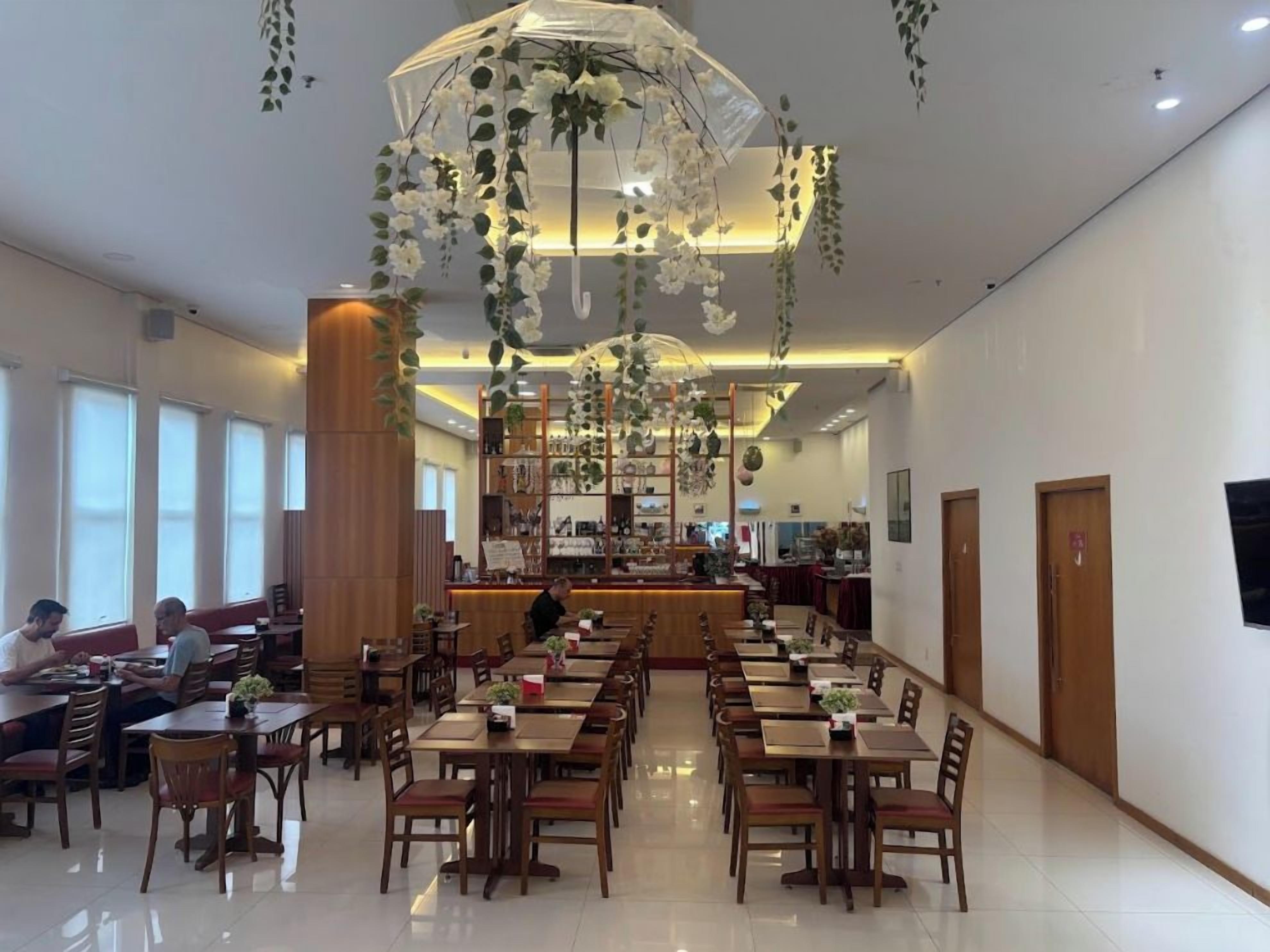 A hotel dining area with multiple tables and chairs, a central bar, and hanging floral decorations.