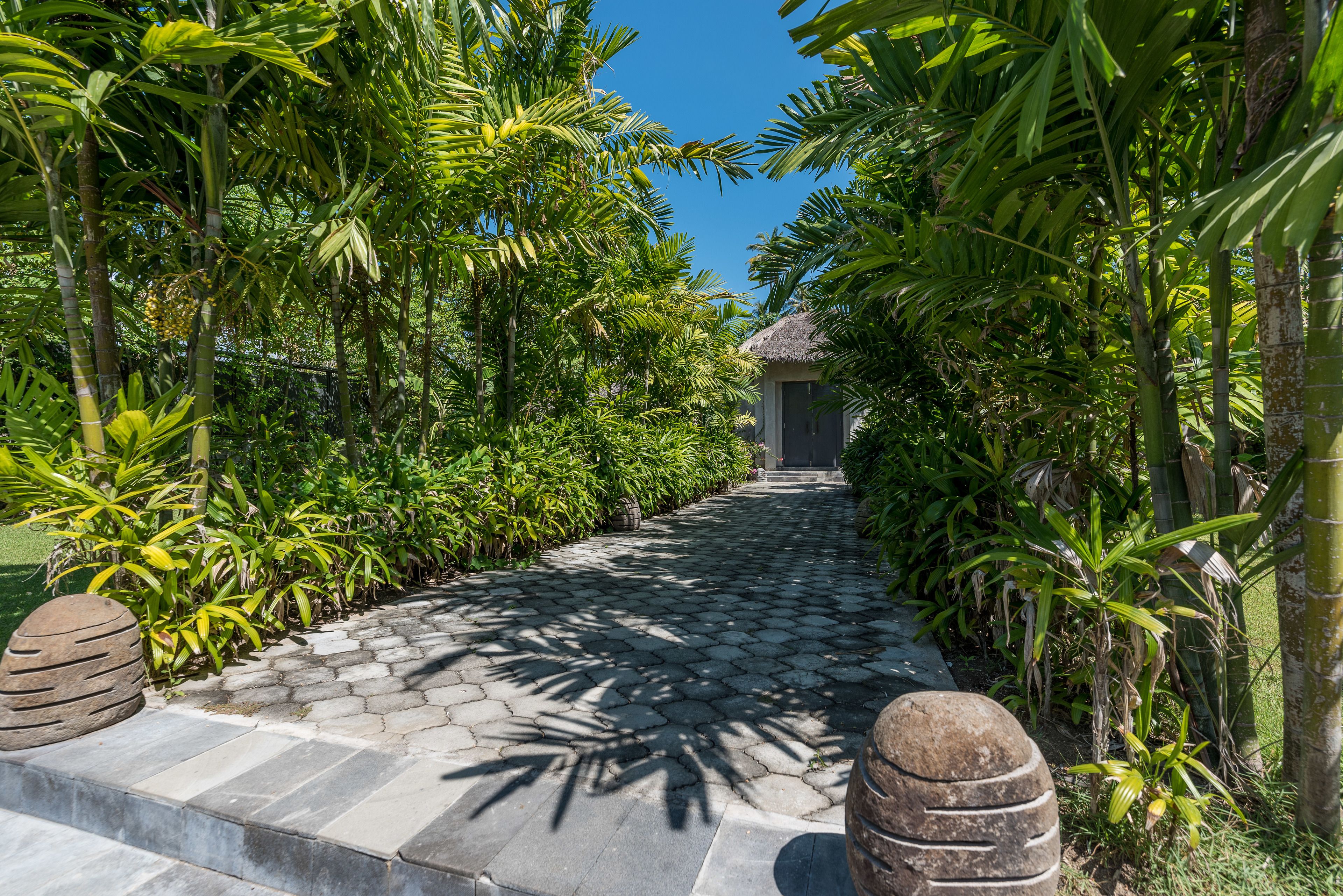 Un sendero pavimentado bordeado por exuberante vegetación tropical que conduce a un edificio con techo de paja.