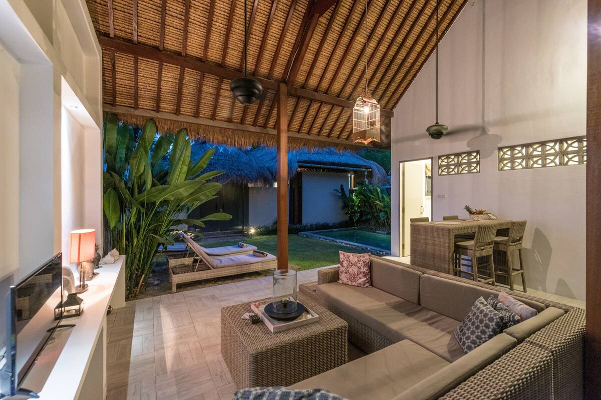 A modern living room with a sofa, a coffee table, and a TV. There is a thatched-roof structure outside, and a dining area with wicker furniture.