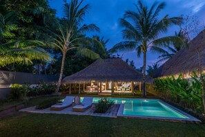 Un espace aménagé au bord de la piscine, avec des chaises longues, une structure à toit de chaume et une piscine entourée de palmiers.