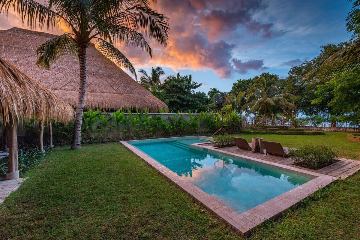 A swimming pool with a thatched roof structure, surrounded by lush greenery and palm trees.