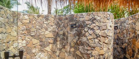 A stone wall with a metal gate, palm trees, and a thatched roof in the background.