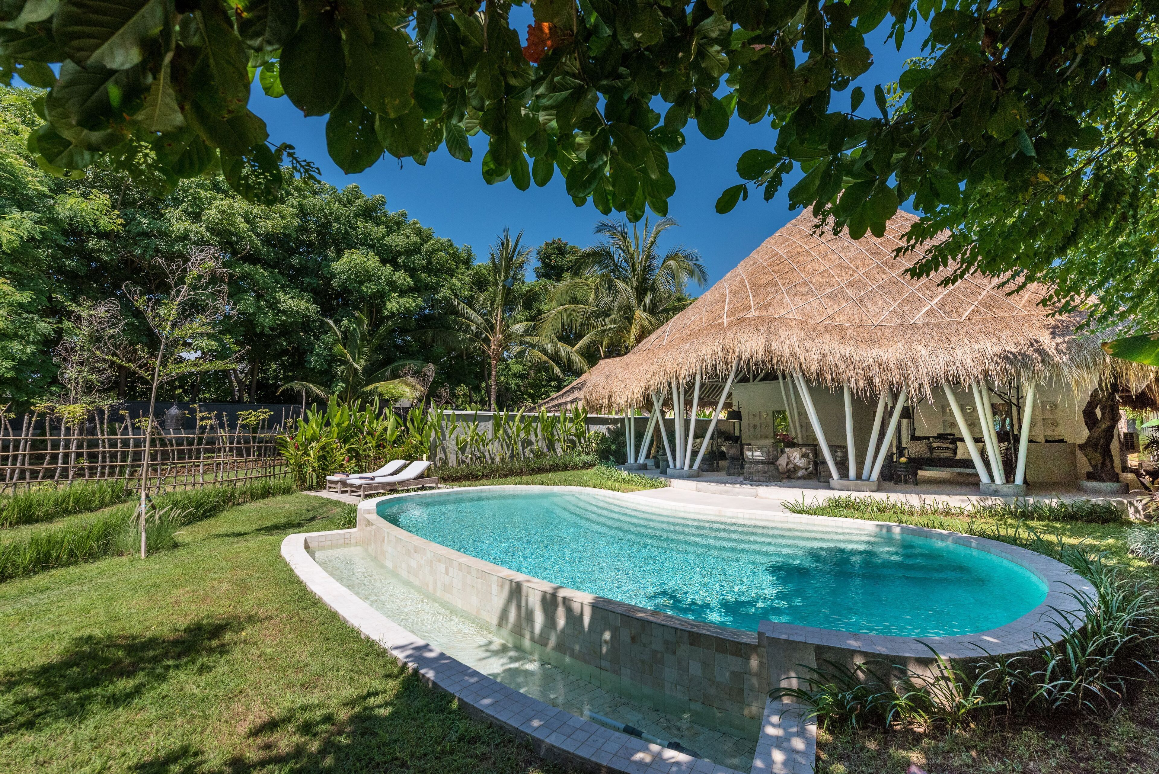 A swimming pool surrounded by lush greenery, a thatched-roof structure, and a fenced area with lounge chairs.