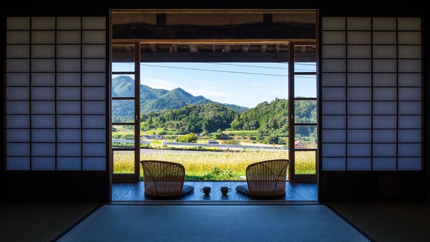 Een traditionele Japanse kamer met tatami vloeren, een lage tafel, twee rieten stoelen en uitzicht op een bergachtig landschap door een shoji scherm.