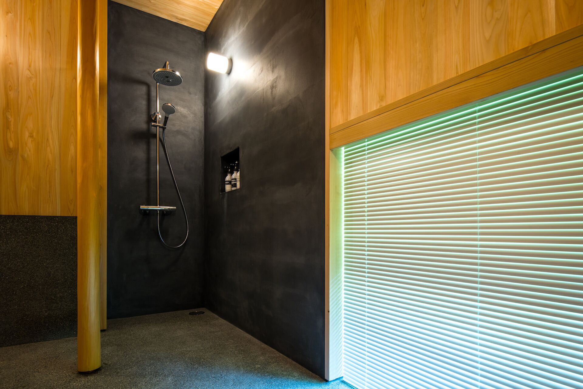 Une salle de bain moderne avec une douche, des murs de couleur sombre, des éléments décoratifs en bois et une fenêtre avec des stores.