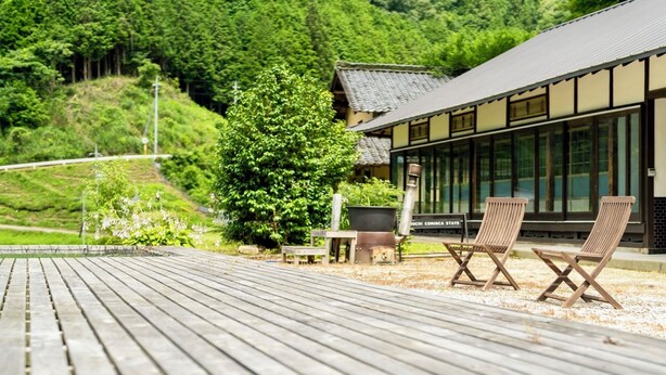 A wooden deck with two chairs and a table, a building with large windows, and a lush green hillside in the background. - Setouchi Cominca Stays Hiroshima FROSEN (Shobara)