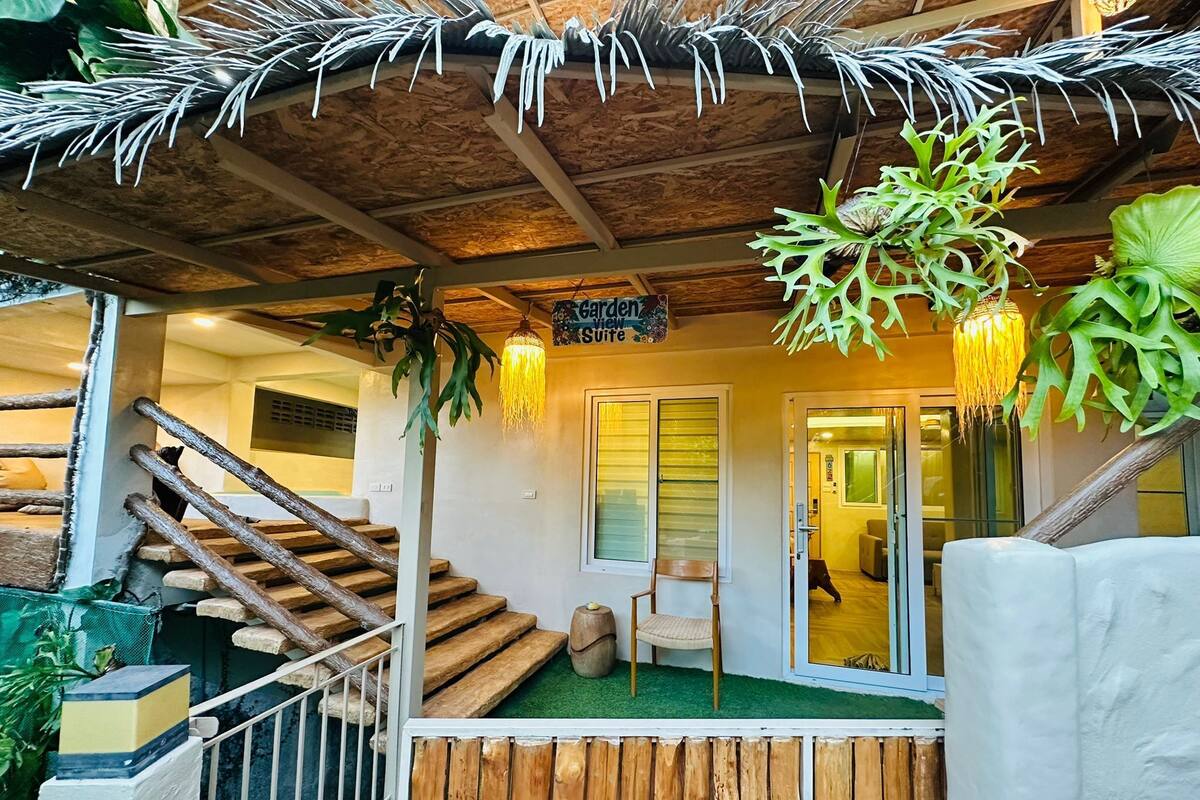Un escalier en bois mène à une pièce dont le toit est recouvert de chaume. Une pancarte indique « Suite avec jardin », et une chaise est placée à l’extérieur.