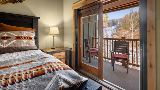 Un dormitorio con cama, mesita de noche, lámpara y vistas a un balcón con sillas.