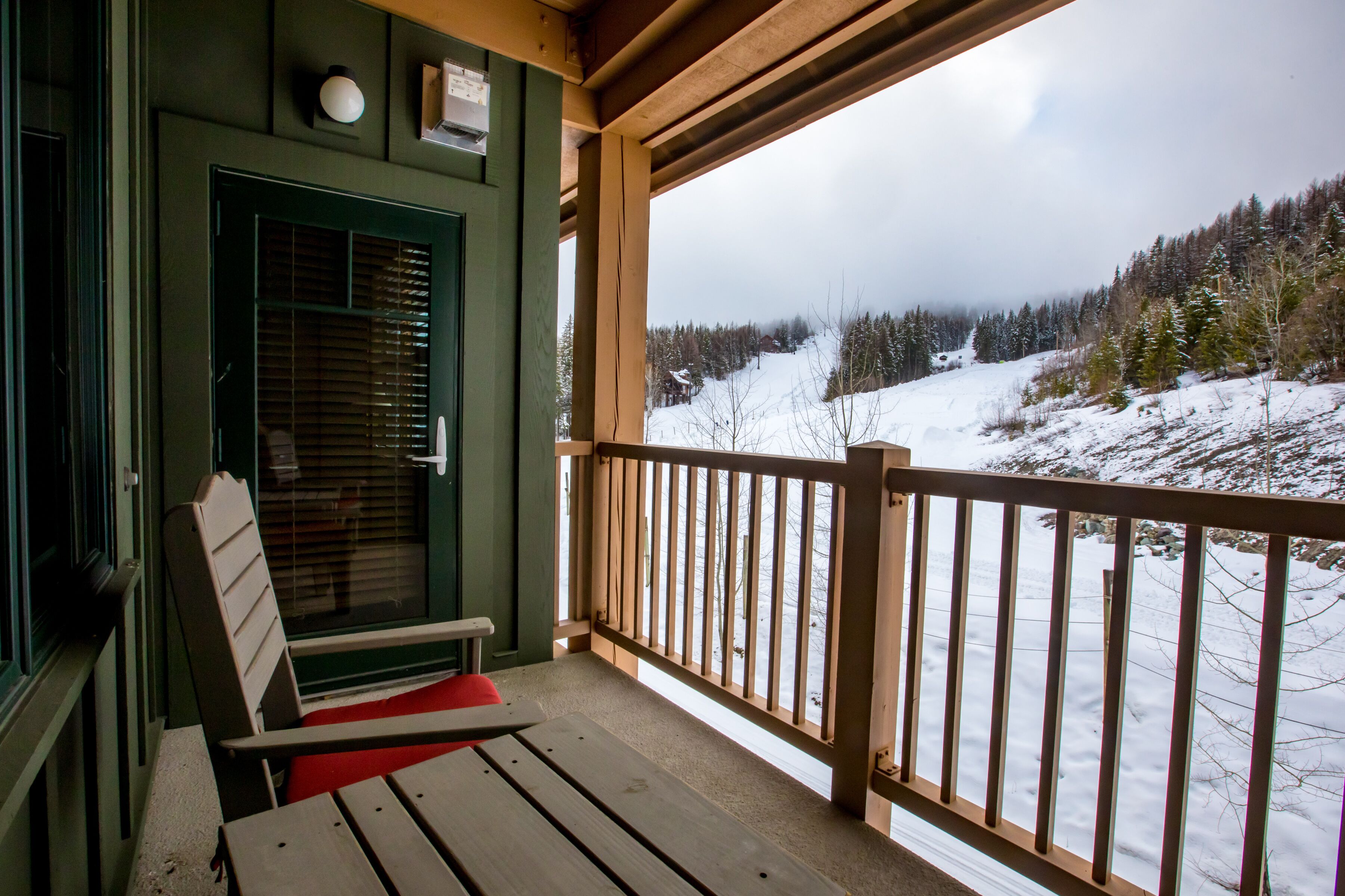 Un terrazza in legno con una sedia rossa e un tavolo, affacciata su un paesaggio innevato con alberi e una montagna all'orizzonte.
