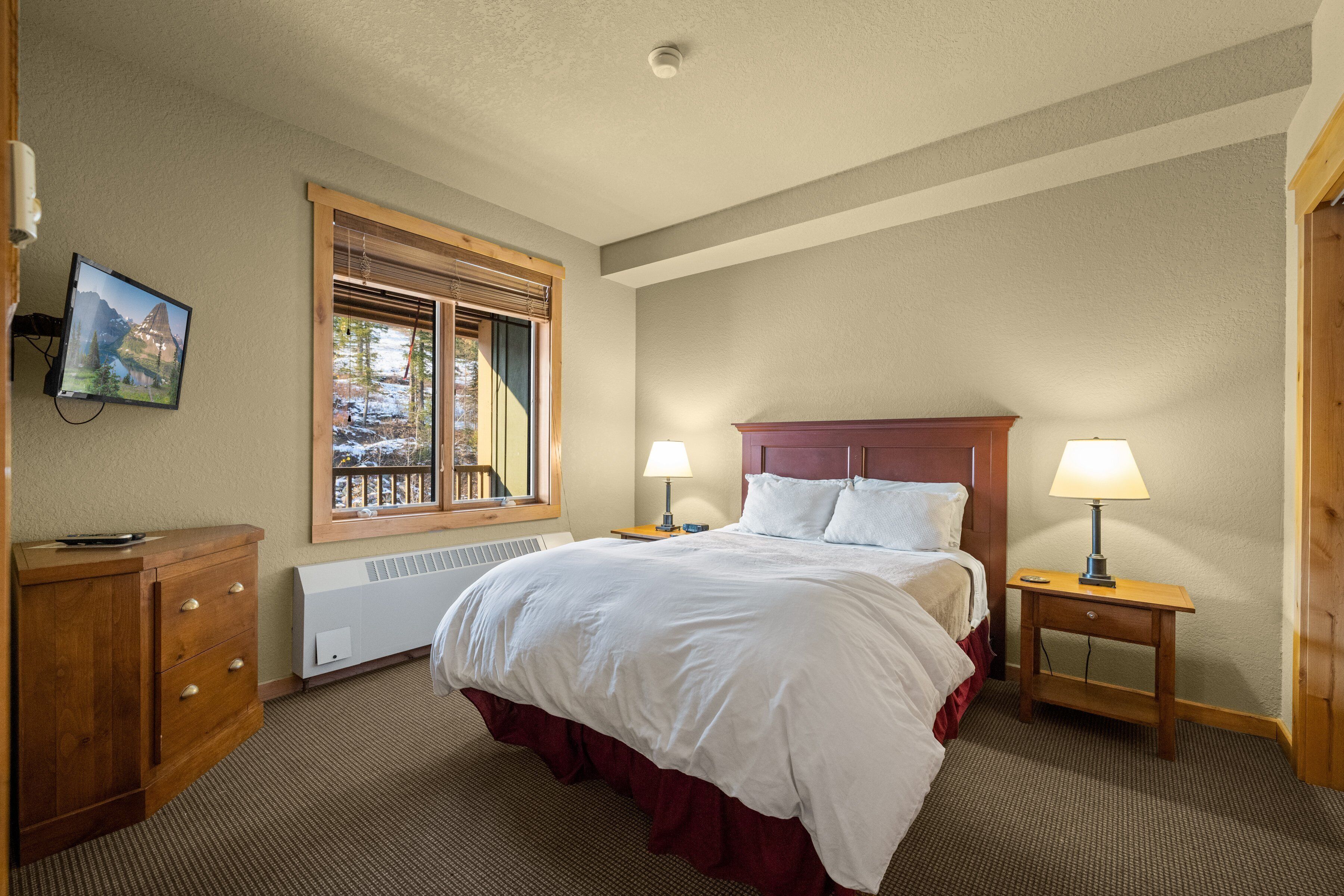 Quarto de hotel com uma cama grande, penteadeira de madeira, janela com persianas, televisão fixada na parede e mesa de cabeceira com abajur.