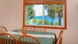 Un coin repas avec une table dressée pour quatre personnes, une vue sur un lac et des arbres par la fenêtre, et une chaise en bois avec un siège en osier.