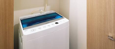 A compact, white, front-loading washing machine with a digital display and control panel, located in a corner of a room with wooden flooring and white walls.