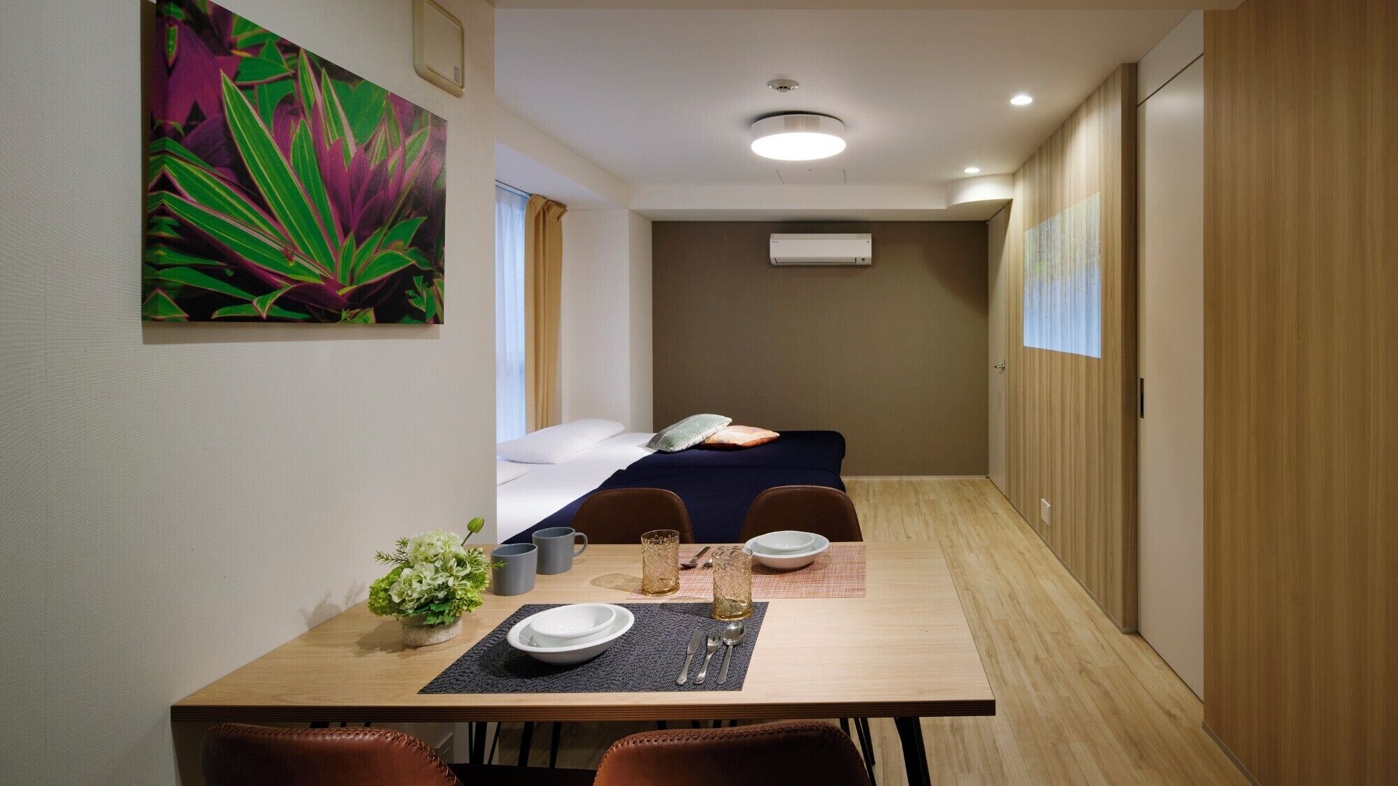 Une chambre d’hôtel de taille modeste, comprenant un lit, une petite table à manger avec des chaises, une plante en pot et un tableau accroché au mur.