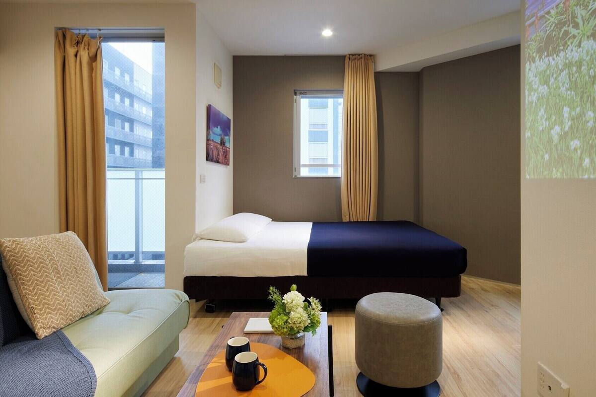 Une chambre d’hôtel avec un lit, un canapé, une table basse avec deux tasses, une petite plante et un écran de projection diffusant une scène de la nature.