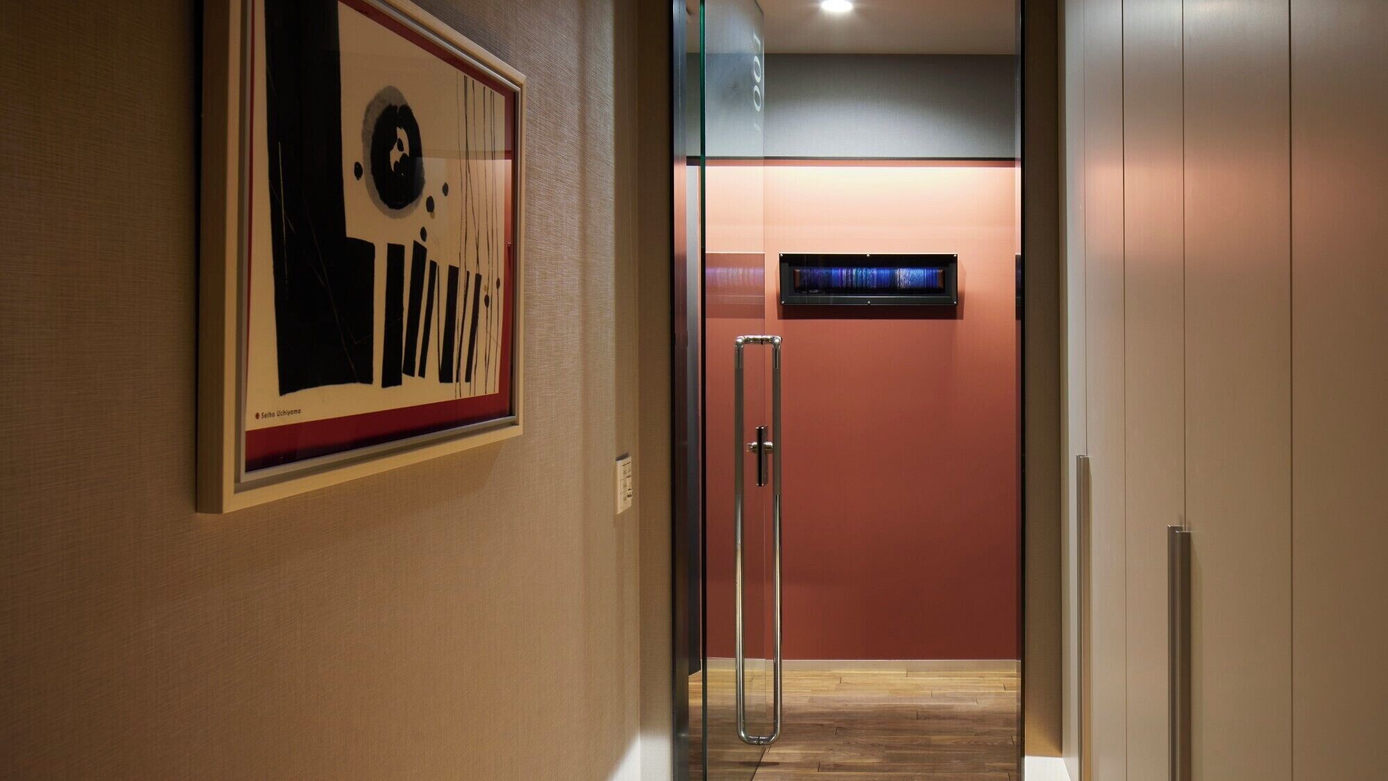 A hallway with a framed abstract artwork, a glass door leading to a room with a red wall and a small window, and a wooden floor.