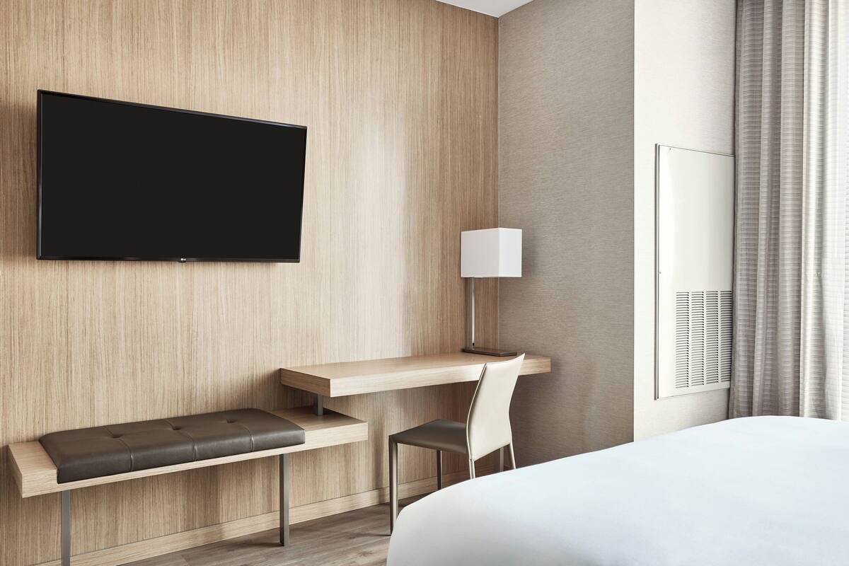 A modern hotel room with a wooden wall, a flat-screen TV, a desk with a chair, a bedside table, and a bed.