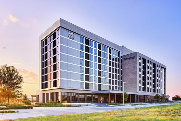 A modern hotel building with a glass facade, a covered entrance, and a landscaped area in front. - AC Hotel by Marriott Dallas Frisco (Frisco)