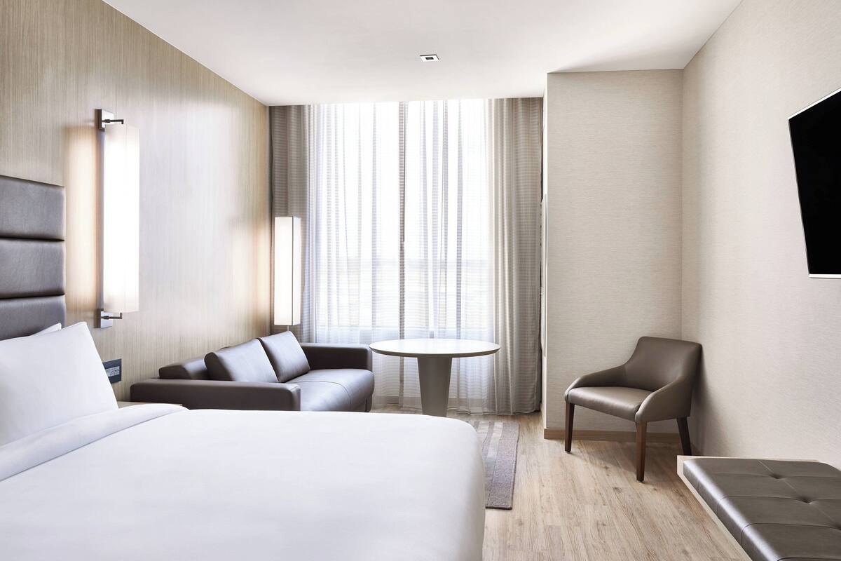 A modern hotel room with a large bed, a sofa, a chair, a small round table, and a TV mounted on the wall.